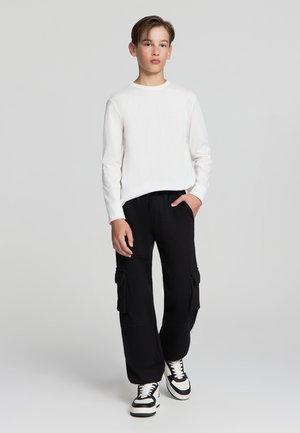 Camicia bianca a maniche lunghe, pantaloni cargo neri con tasche laterali e sneakers bianche e nere. Stile semplice, vestibilità rilassata, texture morbide.