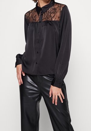 Kobieta ubrana w czarną, długorękawową bluzkę z koronkową wstawką na biuście i satynowym wykończeniem, zestawioną z czarnymi spodniami z ekologicznej skóry.