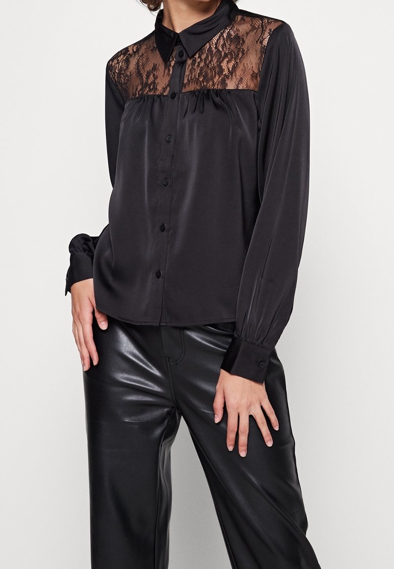 Femme portant un chemisier noir à manches longues avec un yoke en dentelle et une finition satinée, associé à un pantalon en similicuir noir.