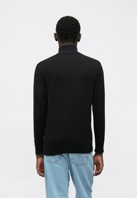 Moški obrnjeno stran v črnem dolgem puloverju s turtleneck ovratnikom in svetlih modrih kavbojkah pred enostavno svetlo ozadjem.