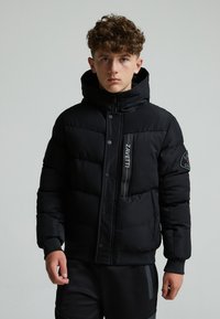 Veste en duvet noire avec un col haut, fermeture éclair et boutons, tissu texturé, et accents de logo sur la poitrine et la manche.