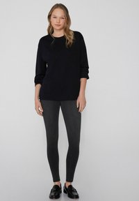 Top negro de manga larga, corte relajado, combinado con jeans ajustados gris oscuro. La modelo lleva zapatos negros sin cordones, de pie contra un fondo gris claro.