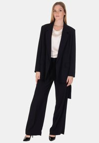 Completo nero composto da un blazer largo con tasche frontali e pantaloni a gamba larga. La blusa è in satin color crema. Le scarpe sono tacchi neri.