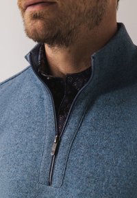 Blue zippered sweater with a textured knit surface, featuring a dark patterned shirt underneath and a stand collar. Silver zipper detail visible.