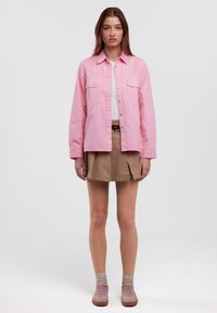Helles rosa Langarmhemd mit Druckknöpfen und Brusttaschen, kombiniert mit einem beigen Faltenrock und pinken Sneakers; neutraler Hintergrund.