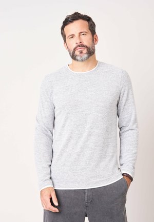 Man met kort donker haar en baard, die een lichtgrijs overhemd met lange mouwen en een donkere broek draagt, staande tegen een effen lichte achtergrond.