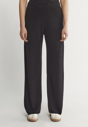 Mujer con pantalones negros sueltos y zapatillas blancas, de pie contra un fondo claro y sencillo.