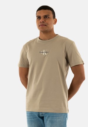 T-shirt in cotone beige con collo a giro e maniche corte, caratterizzata da un logo "Calvin Klein Jeans" in azzurro chiaro sul petto.