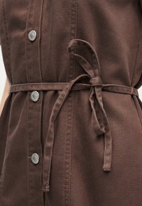 Καφέ φόρεμα από ύφασμα με ασημί κουμπιά κλιπ και ζώνη στη μέση, που αναδεικνύει τις λεπτομέρειες υφής και ραφής.