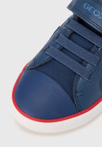 Kék sportcipő sima bőr orrmerevítővel, texturált szövet díszítéssel, tépőzáras pánttal és piros gumitalppal. A fehér középtalp kontrasztot ad.