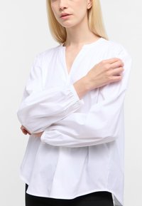 Hvid langærmet bluse med V-hals, samlede manchetter og en afslappet pasform. Glat tekstur og subtil glans med en buet nederste kant.