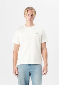 T-shirt en coton crème avec des manches courtes, un col rond et un petit logo brodé sur la poitrine gauche. Associé à un jean en denim bleu clair.