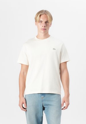 Cream-colored cotton t-shirt with short sleeves, round neckline, and small embroidered logo on the left chest. Paired with light blue denim jeans.