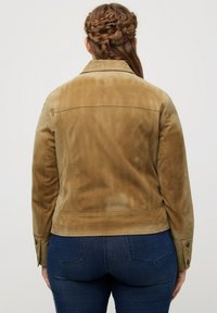 Tan mockaskinnjacka med en figurnära design, tryckknappdetaljer vid ärmarna och subtil textur. Bärs över mörkblå jeans.