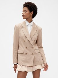 Blazer beige à double boutonnage avec des revers crant�és, deux poches à l'avant et des boutons marron ; porté sur une chemise blanche et une jupe superposée.