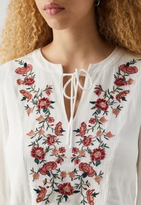 Vit broderad blus med röda och gröna blommönster, V-ringning med knytning och en texturerad tyg.