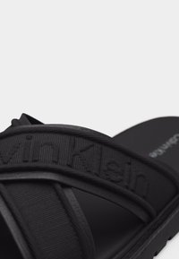 Sandale noire avec des larges brides croisées en tissu embossées du logo "Calvin Klein" sur un fond clair uni.