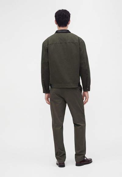 Homme debout, de dos, portant une veste vert foncé et un pantalon assorti avec des chaussures foncées sur fond blanc uni.