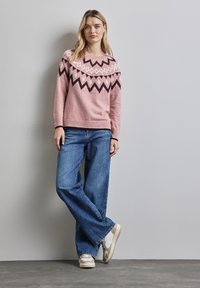 Ljusrosa tröja med geometriska mönster i mörkbrunt och grädde. Kombinerad med utsvängda blå jeans och sneakers i grädde. Enkla designelement.