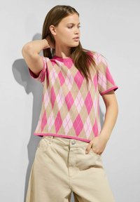 Krátký rukáv svetr má růžovo-béžový argyle vzor, s žebrovaným kulatým výstřihem a kontrastním lemem. V kombinaci s béžovými kalhotami.