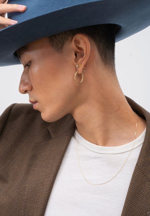 Blauer Filzhut, goldene Ringohrringe, goldene Stecker, brauner strukturiert Blazer, weißes T-Shirt, minimalistisches goldene Halskette. Seitenansicht.