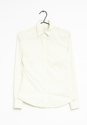 Chemise boutonnée jaune clair avec des rayures blanches verticales, manches longues, col pointu et ourlet droit. Accrochée à un crochet noir.