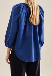 Camicia a maniche lunghe di un blu profondo, con scollatura arricciata, tessuto testurizzato e orlo arrotondato, abbinata a pantaloni scuri.