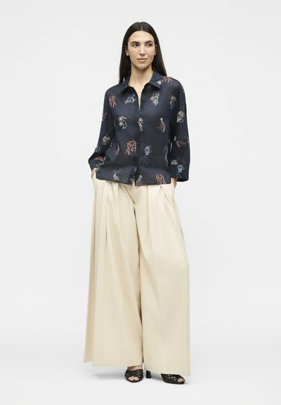 Femme aux longs cheveux foncés portant un chemisier bleu marine à motifs et un pantalon large beige, debout les mains dans les poches sur fond blanc.