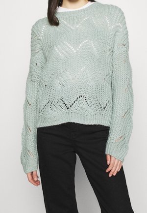 Pull en tricot teal clair à coupe décontractée, doté d'un motif en vagues, d'une texture ajourée et de manches longues. Associé à un pantalon noir.