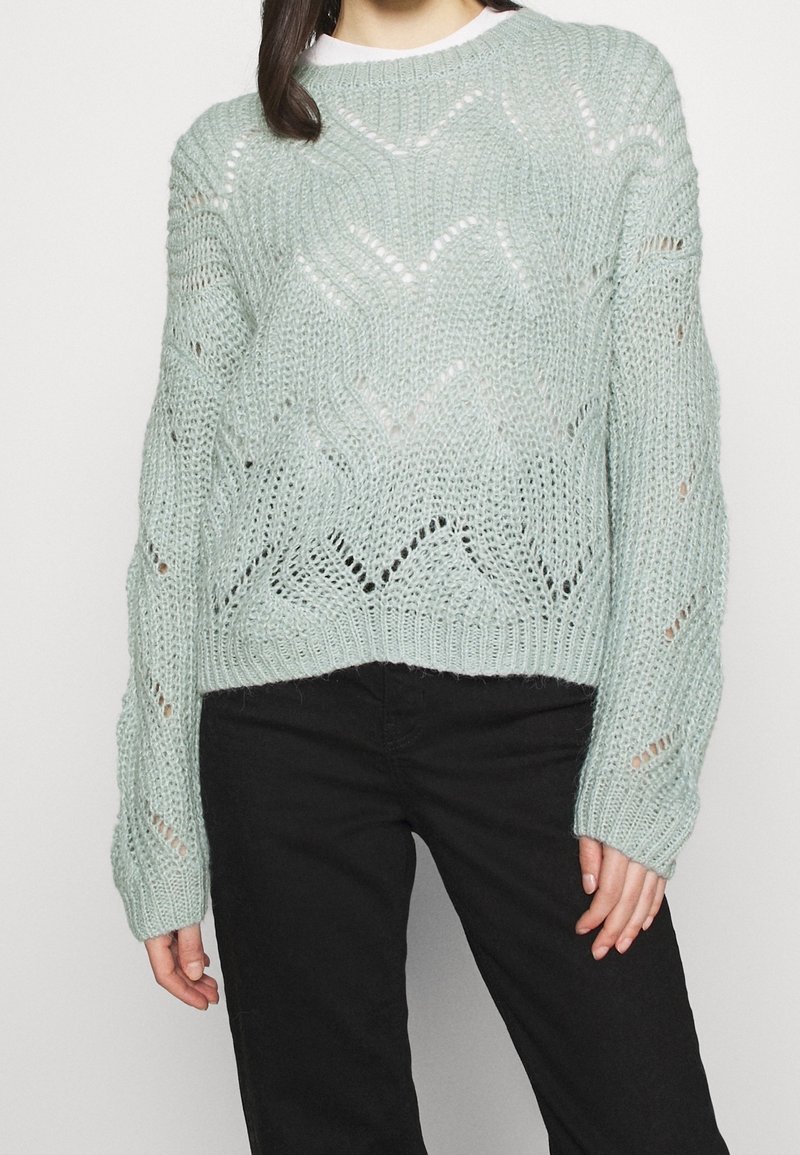 Pull en tricot teal clair à coupe décontractée, doté d'un motif en vagues, d'une texture ajourée et de manches longues. Associé à un pantalon noir.