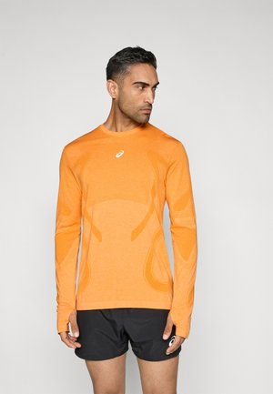 Oranžové atletické tričko s dlhým rukávom z textúrovaného materiálu, s kontrastnými detailmi a otvormi na palce. Kombinované s čiernymi šortkami.