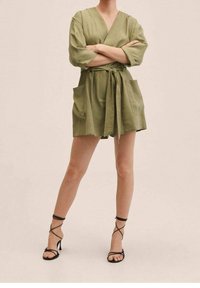 Vestido cruzado verde oliva con escote en V, mangas cortas, dos bolsillos delanteros y cintura con cinturón, combinado con sandalias negras de tacón alto con tiras.