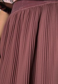 Plisserad kjol i dämpad mauve med en slät, texturerad midja. Har flödande veck och ett mjukt, lätt material.