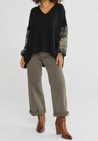 Femme portant un pull noir à col en V avec des manches transparentes à motifs, un pantalon large olive retroussé et des bottines marron à talons.