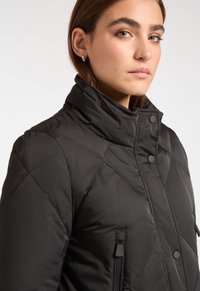 Jonge vrouw met lichte ogen, gekleed in een zwarte gewatteerde jas met hoge kraag en ritszakken, die iets naar de zijkant kijkt.