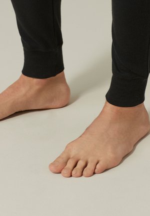Pieds nus portant un pantalon noir ajusté à la cheville, debout sur un sol clair.