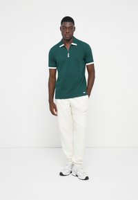 Grøn polo skjorte med lynlåskrave, hvidt kantbånd på ærmerne, parret med cremefarvede joggingbukser og grå sneakers. Enkelt design, stof i bomuldsblanding.