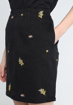 Zwarte rok met kleine gele en roze geborduurde bloemen, gedragen met één hand in de zak en zwarte top.