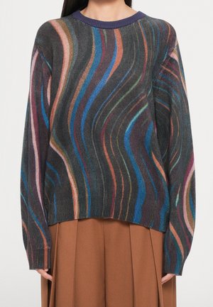 Kobieta ubrana w wielokolorowy, falisty sweter w paski w odcieniach niebieskiego, pomarańczowego, różowego, zielonego i czarnego oraz brązową plisowaną spódnicę.