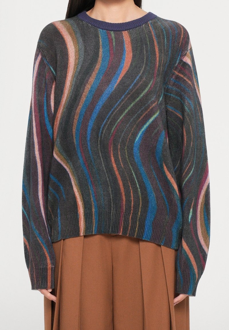 Femme portant un pull multicolore à rayures ondulées avec des tons bleus, orange, rose, vert et noir, ainsi qu'une jupe plissée marron.