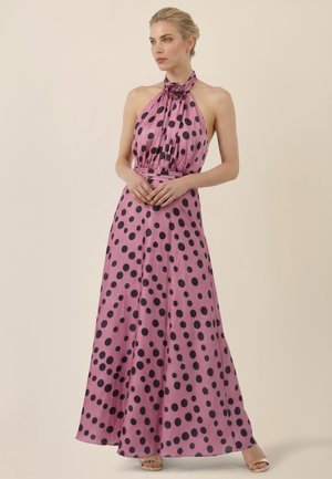 Frau trägt ärmelloses rosa Neckholderkleid mit großen schwarzen Punkten, floraler Halsdetail und beigen offenen Sandalen, steht vor einem einfarbigen Hintergrund.