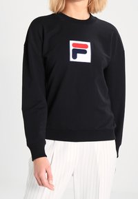 Persona vistiendo un sudadera negra con un parche de logo rojo y azul marino en el pecho y pantalones blancos a rayas.