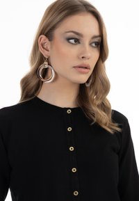 Maglietta nera a maniche lunghe con scollatura rotonda e bottoni dorati. Orecchini circolari grandi con accenti trasparenti e marroni.