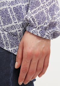 Handen vilar vid sidan i en långärmad blus med blåa blommönster och mörkblå denimbyxor.