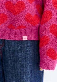 Pletený růžový svetr s červenými srdíčkovými vzory, žebrovanými manžetami a malou růžovou visačkou značky. V kombinaci s tmavými džíny s červeným prošíváním.