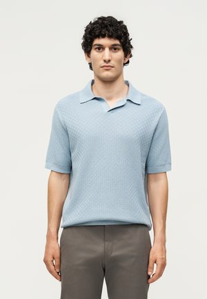 Homme aux cheveux courts, bouclés et noirs, portant un polo bleu clair texturé à manches courtes et un pantalon gris, face à un fond uni.