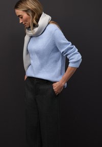 Vaaleansininen resorineule, jossa on timantinmuotoinen kuvio, yhdistetty tummanharmaisiin leveälahkeisiin housuihin. Pehmeä harmaa huivi laskeutuu kaulan ympärille.