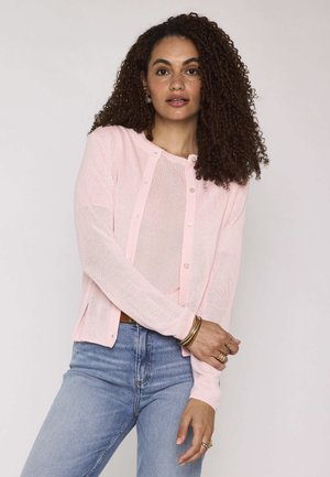 Donna con capelli ricci che indossa un cardigan rosa chiaro con bottoni sopra una maglietta rosa abbinata e jeans blu, accessoriata con braccialetti e anelli d’oro.