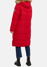 Doudoune rouge avec une capuche bordée de fourrure, présentant des doublures horizontales et un design slim. La personne porte un jean bleu et des bottes noires.