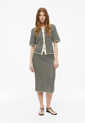 Femme aux longs cheveux roux portant un gilet à manches courtes rayé noir et blanc et une jupe midi assortie avec des sandales noires.